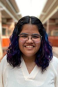 Woman with olive skin and shoulder length curly black hair with blue and purple pieces wearing a whiteshirt and glasses